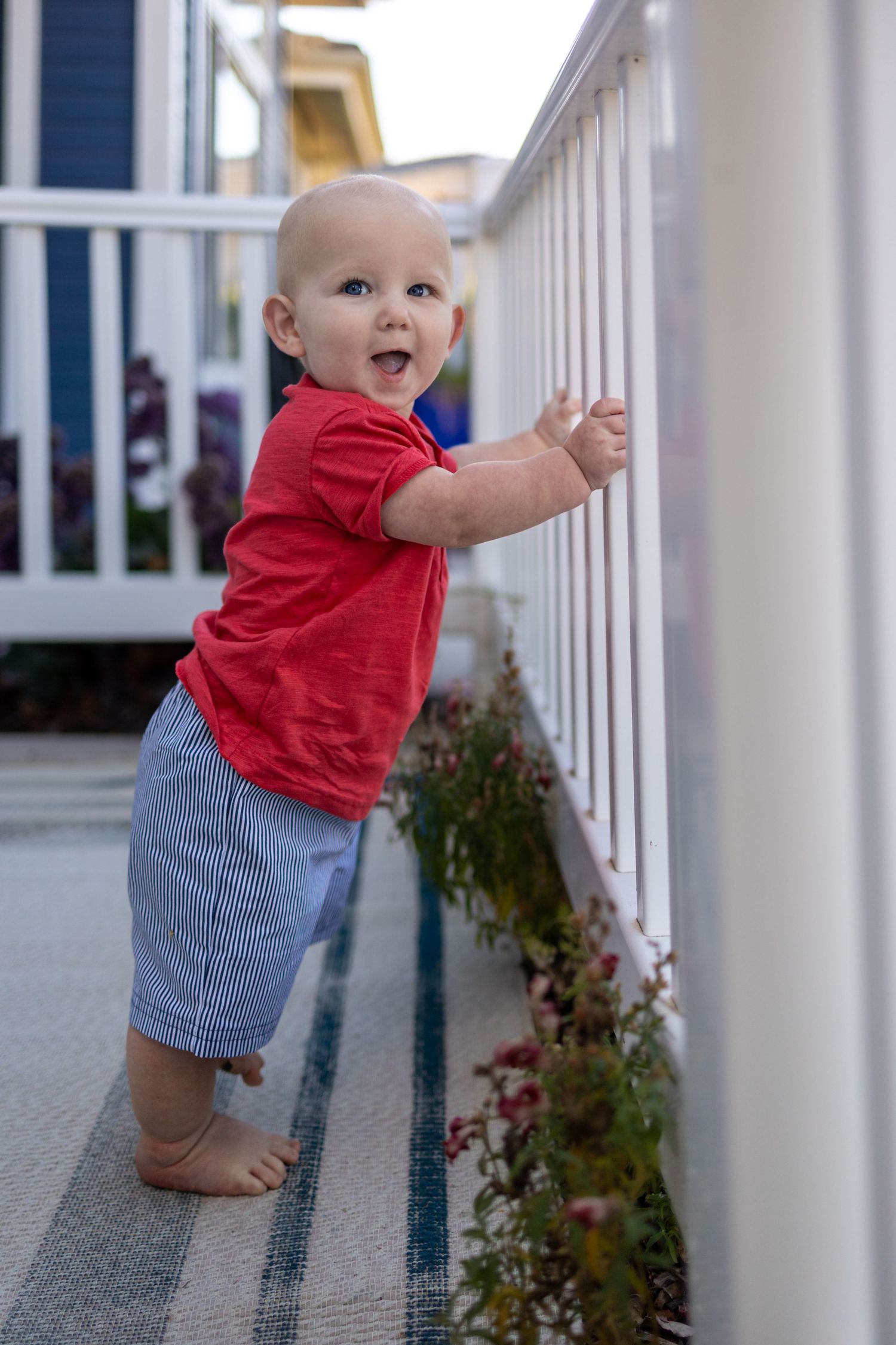 Baby in red shirt and striped shorts standing at white porch railing.
