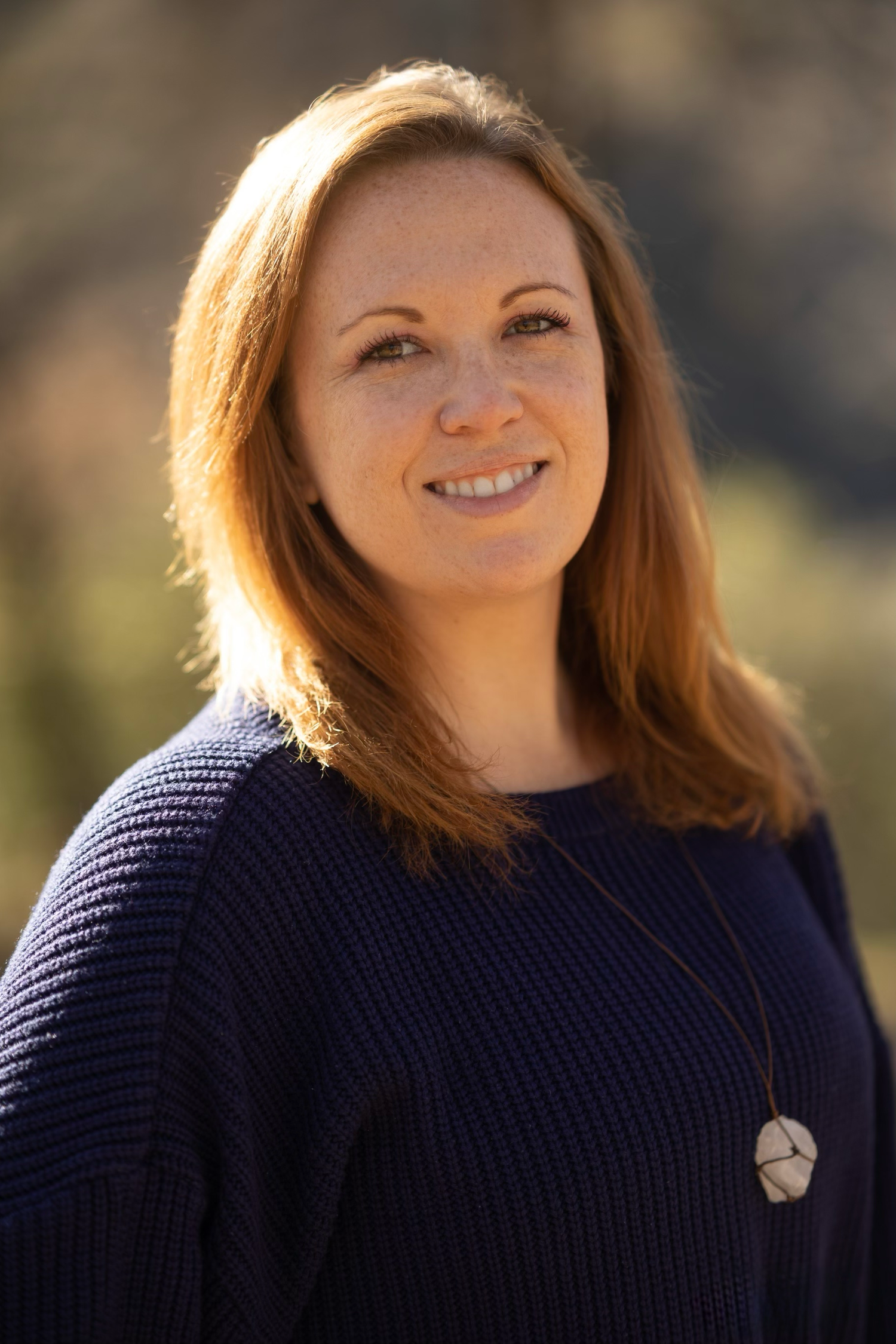 Professional headshot portraits in a navy sweater against a blurred outdoor background.