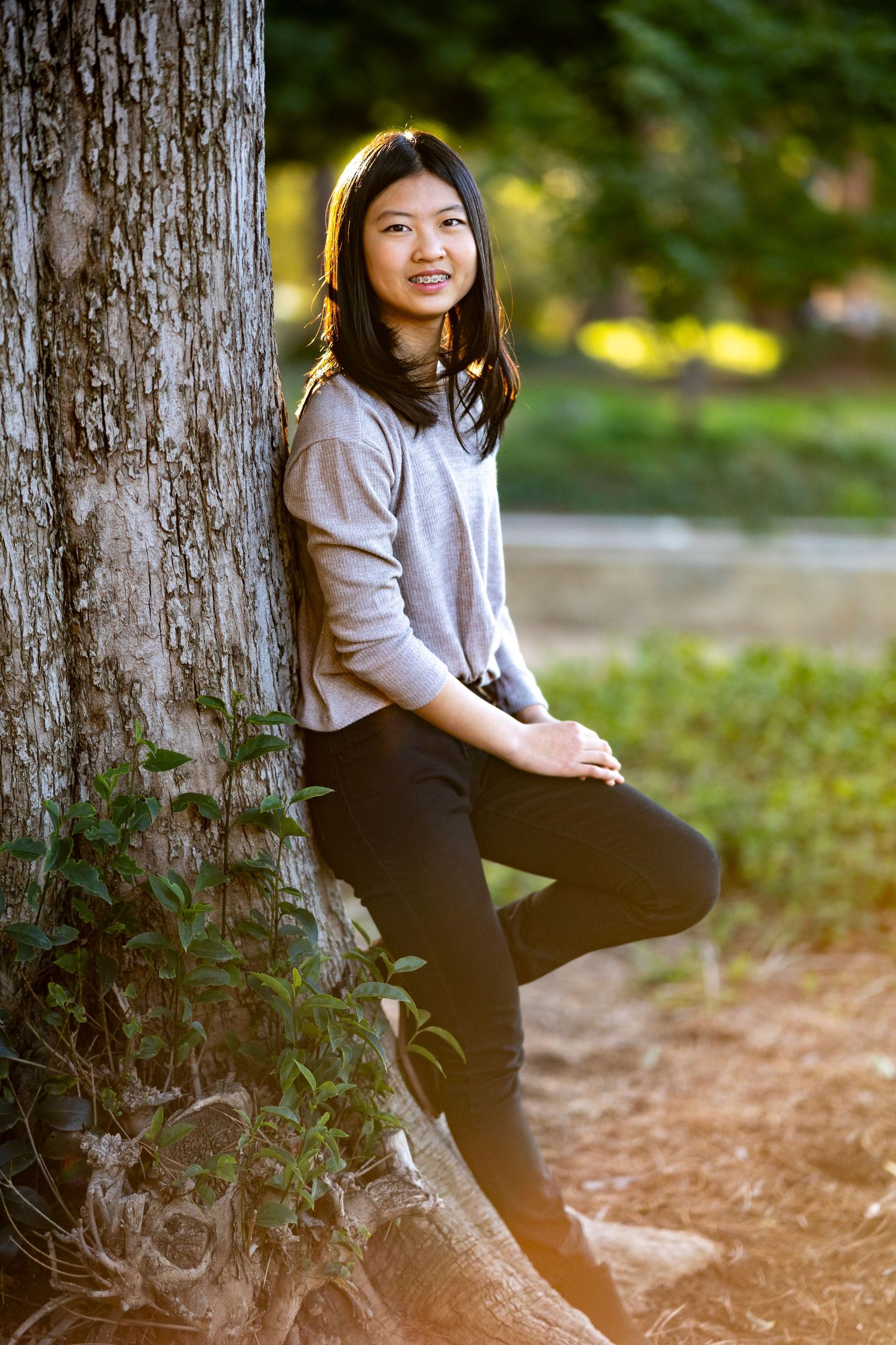 Person in casual sweater leans against large tree trunk in outdoor setting.