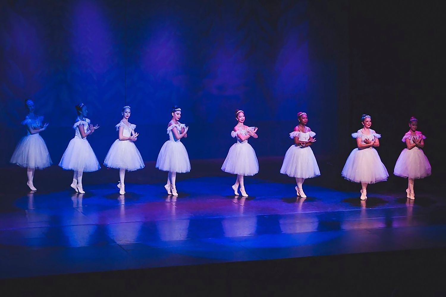 Ballet dancers in white tutus performing in formation on a blue-lit stage during a classical dance performance.