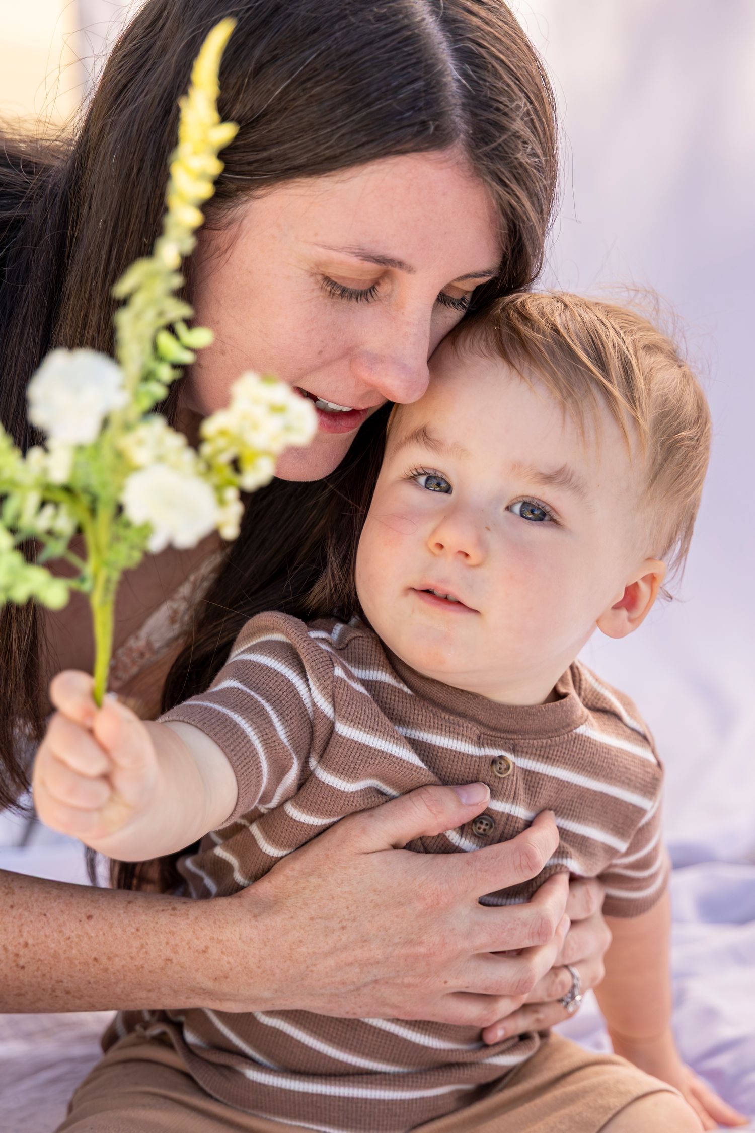 A loving mother holds her baby boy while he clutches a small white flower bouquet outdoors.