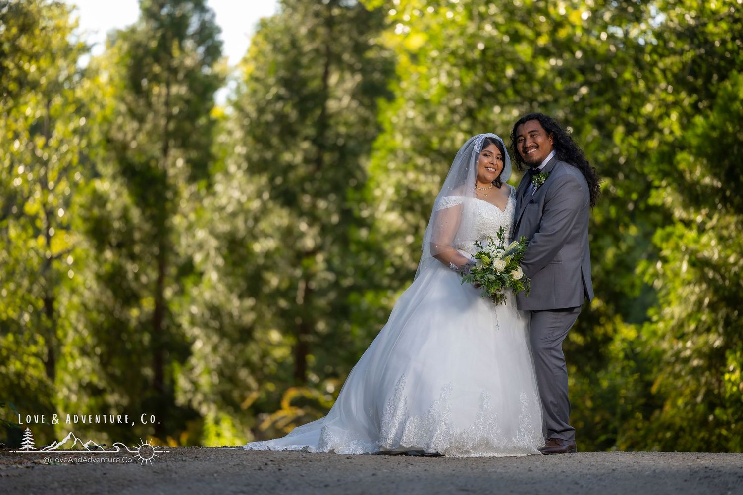 A couple in wedding attire poses for portraits in a lush green outdoor garden setting.