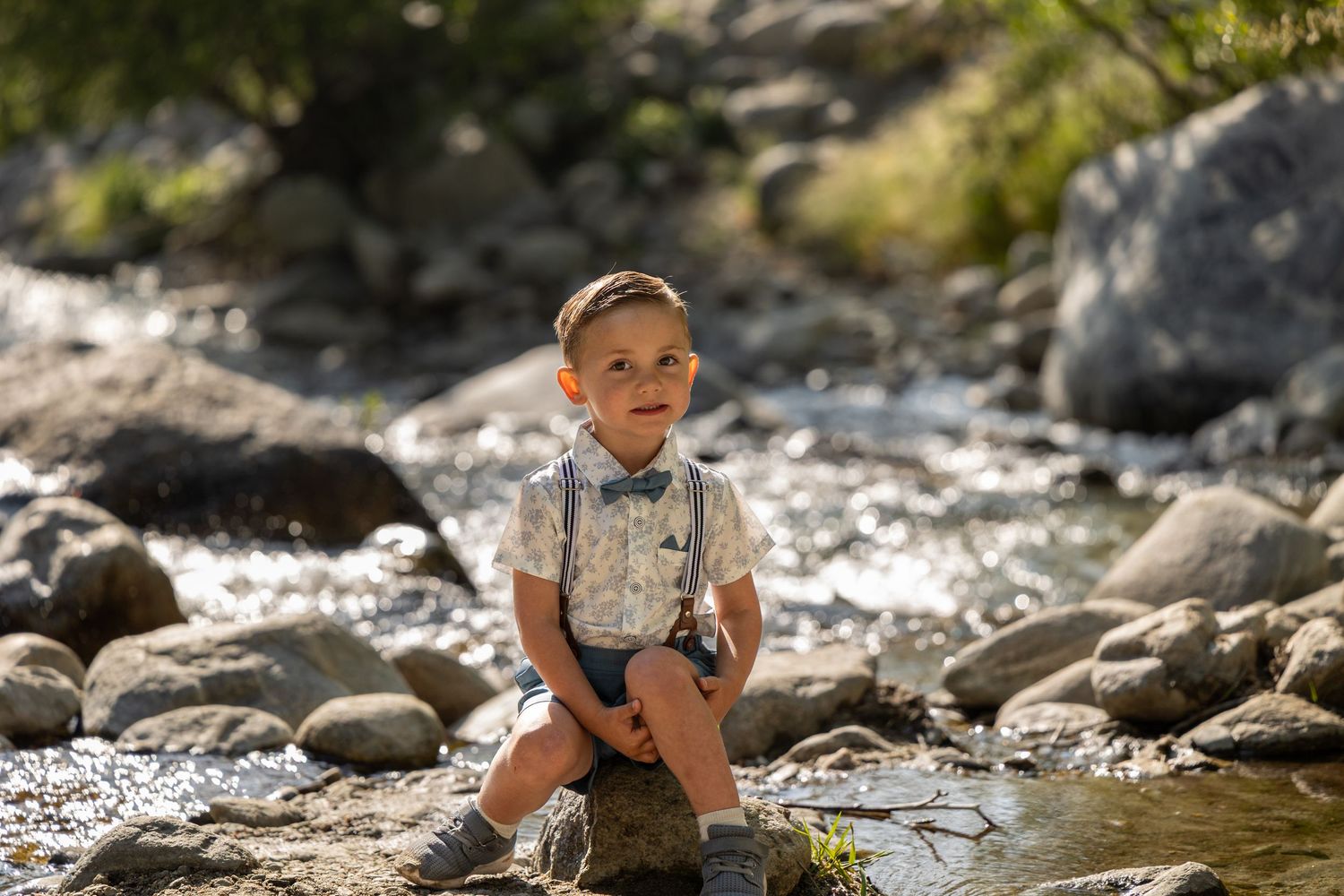 Young child in suspenders sitting on rock in mountain stream.