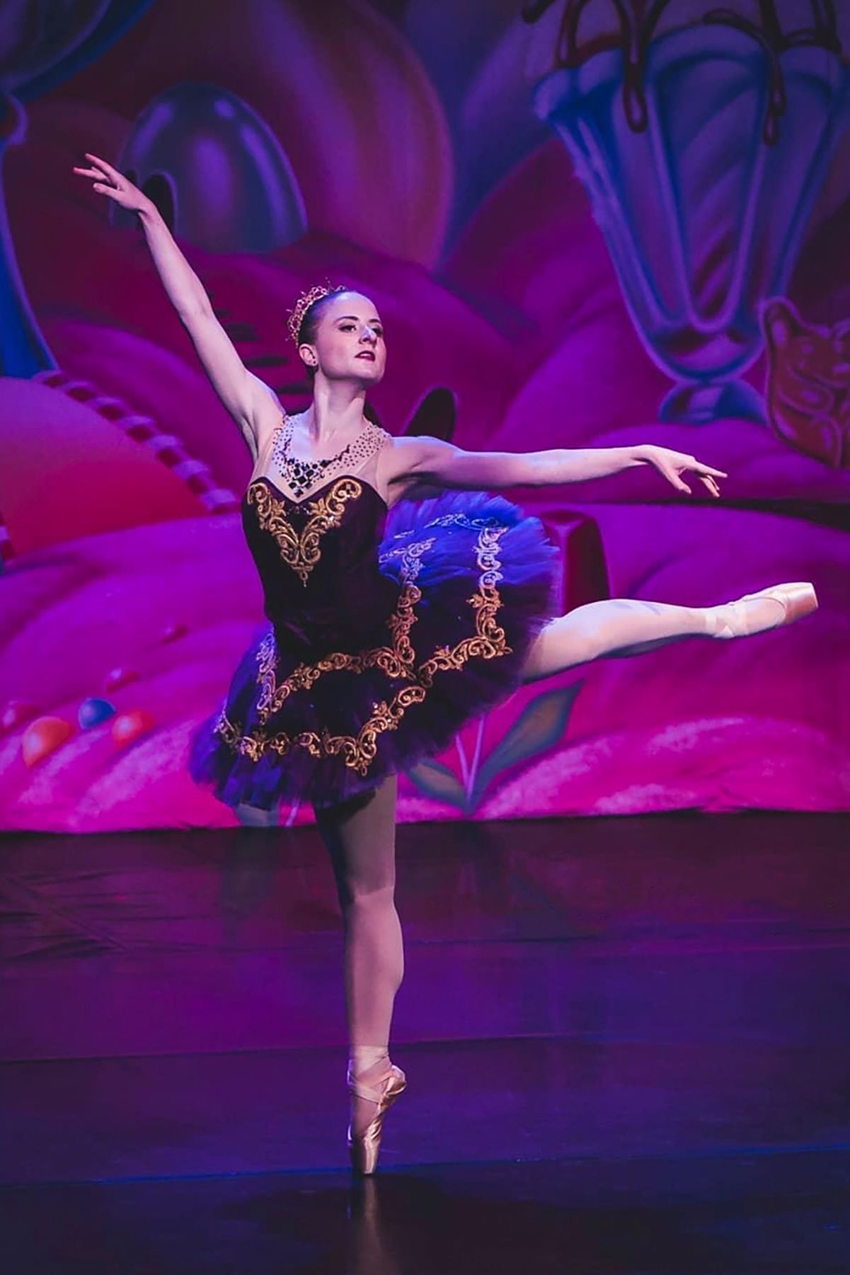 Ballerina in black and gold tutu performing an arabesque pose against colorful stage backdrop.