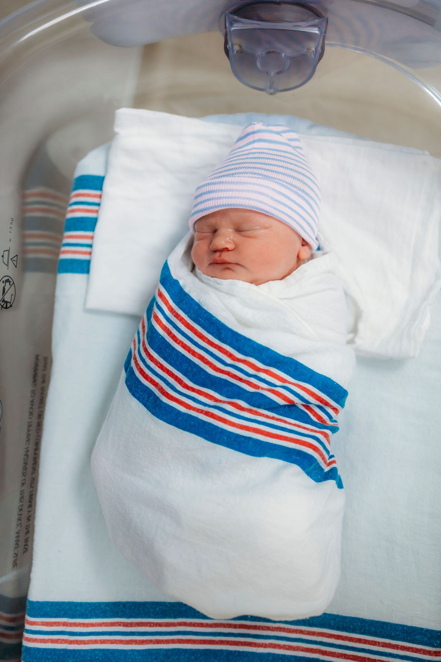 Sleeping newborn baby wrapped in white hospital blanket with blue and red stripes while resting in bassinet.