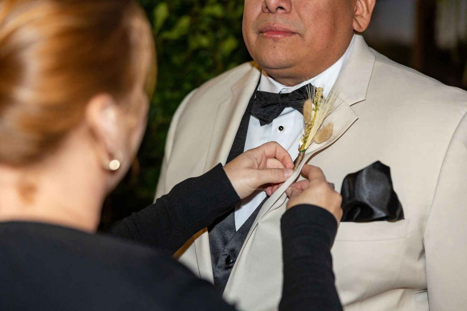 Someone pins a white floral boutonniere onto a cream colored tuxedo jacket lapel during a formal event.