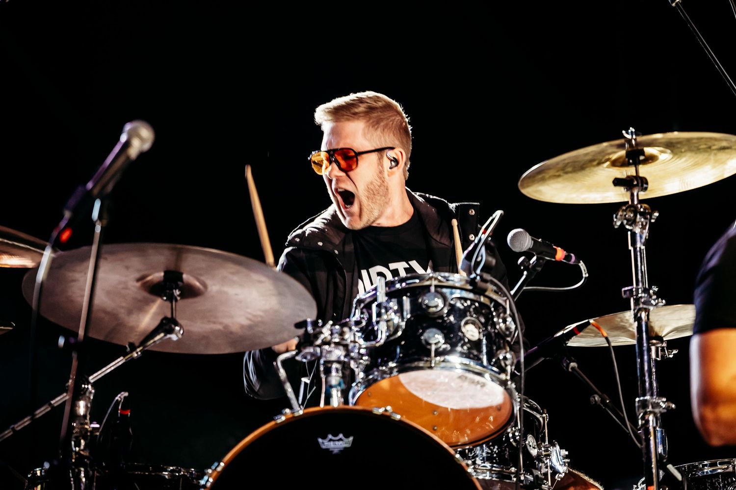 Energetic drummer with glasses performing behind drum kit during concert.
