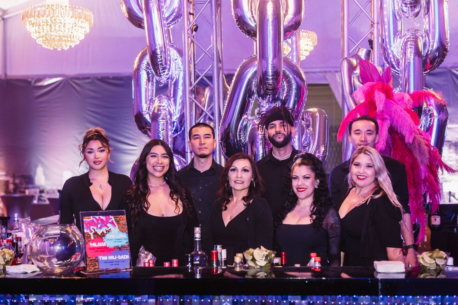Performers in black outfits sit at tables during a stage production with purple lighting and decorative elements.