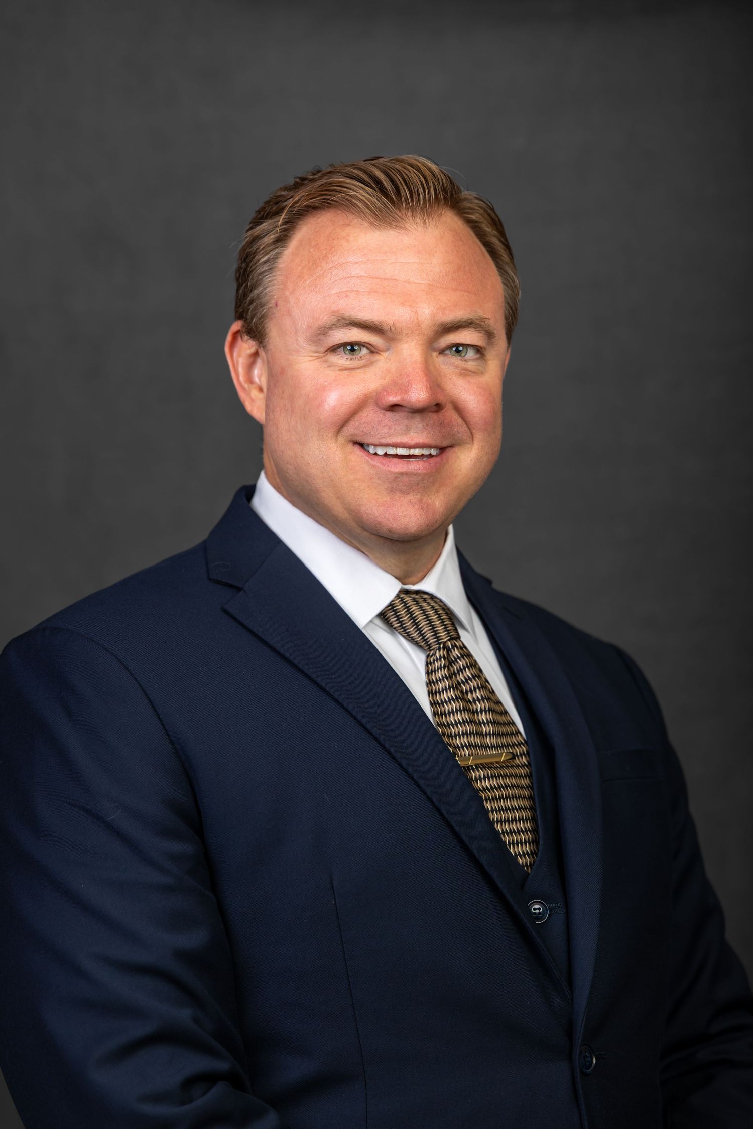 Professional headshot of someone in a navy suit and striped tie against a dark background.