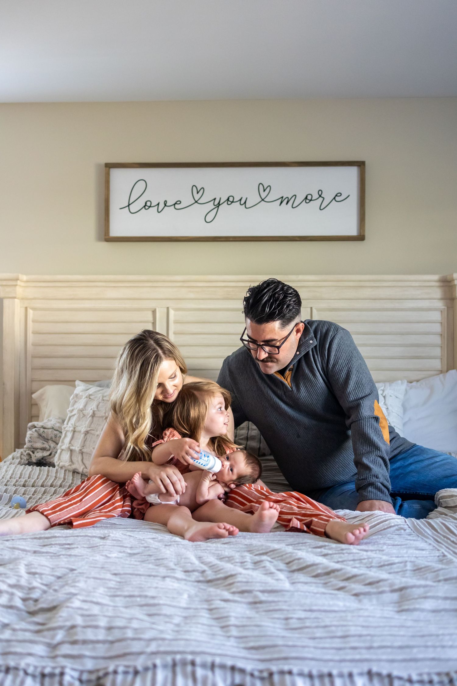 Family scene on bed with 'love you more' sign visible on wall.