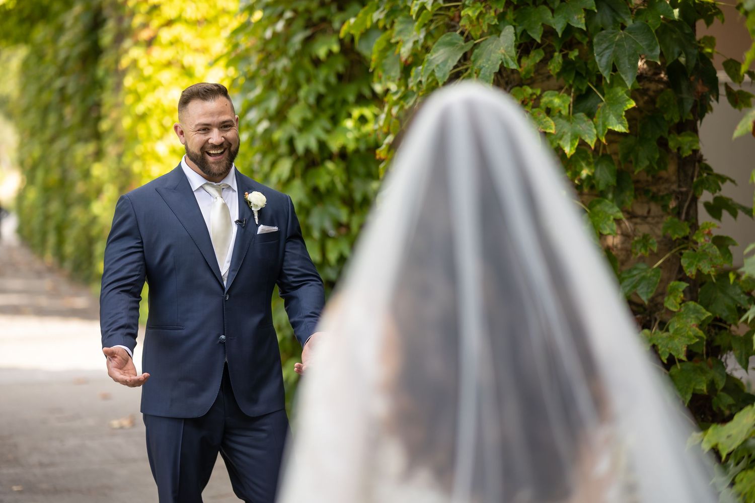 A joyful first look wedding moment between a groom in a navy suit and bride in a flowing white veil outdoors.