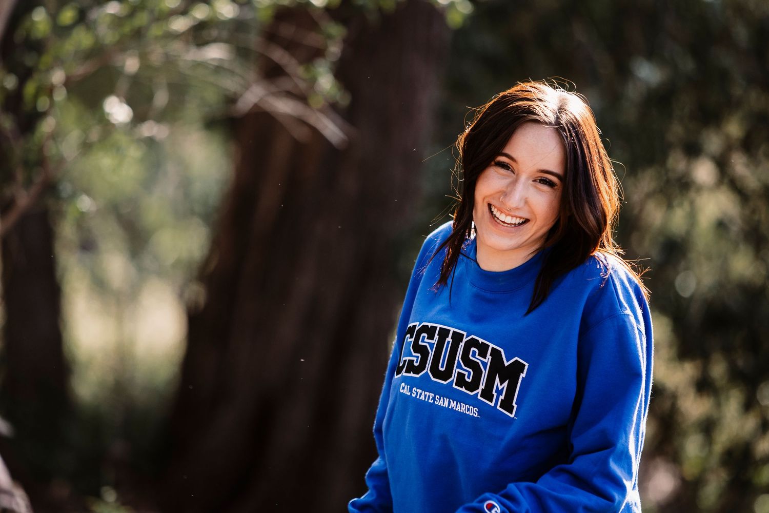 Graduate a bright blue university sweatshirt smiles outdoors against a natural backdrop in Redlands.