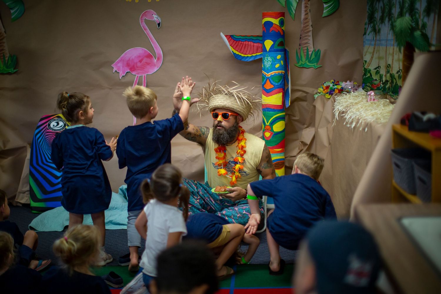 Students in blue uniforms gather around a classroom display featuring colorful tropical decorations including a flamingo and parrot.