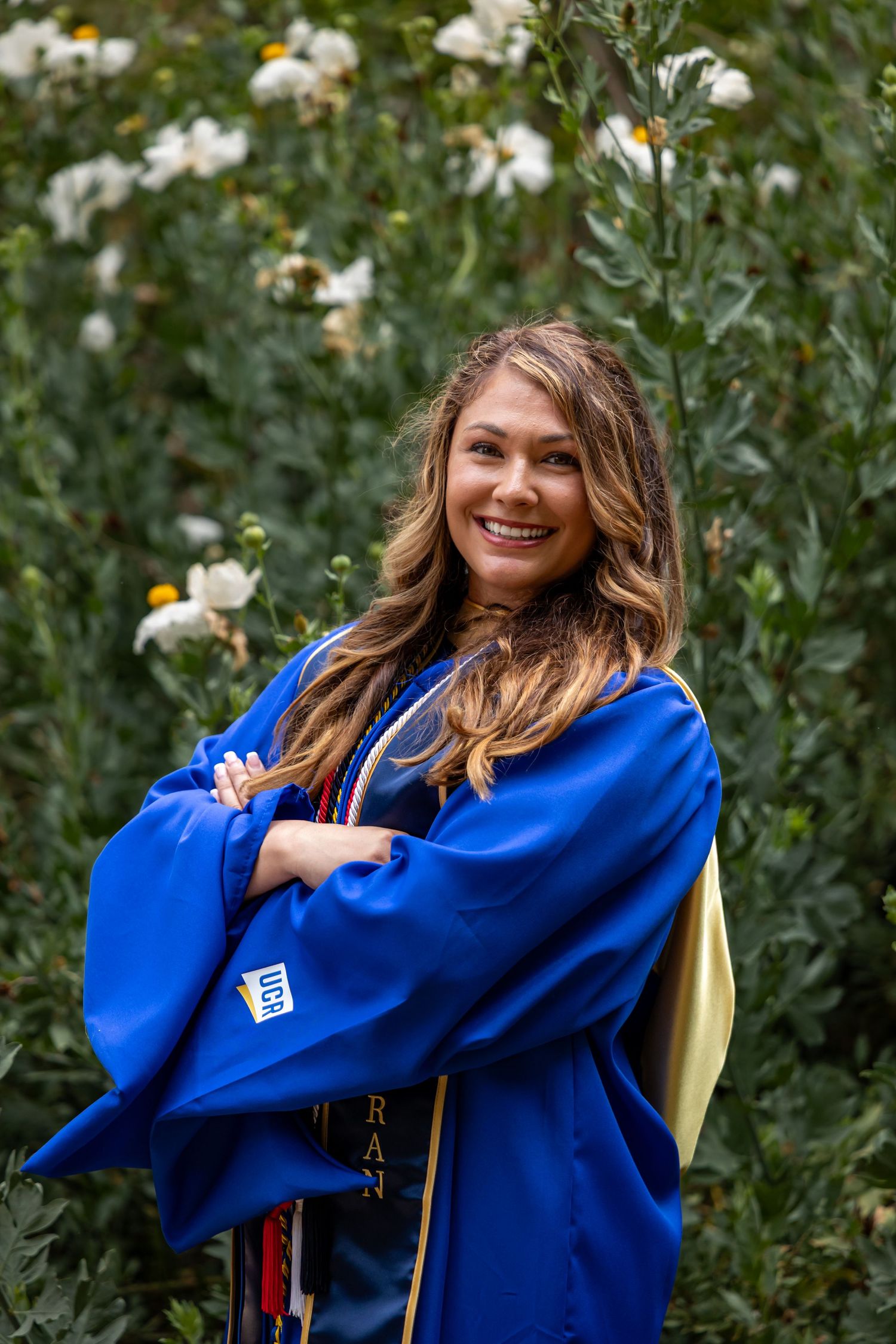 UCR graduate wearing blue commencement regalia poses in garden with white flowers at University of Riverside Botanical Gardens.