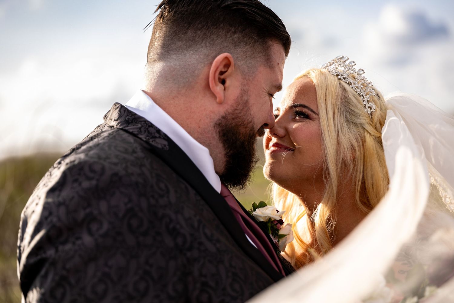 Wedding couple embraces while bride's veil blows in the wind against a soft cloudy sky backdrop in Fullerton.