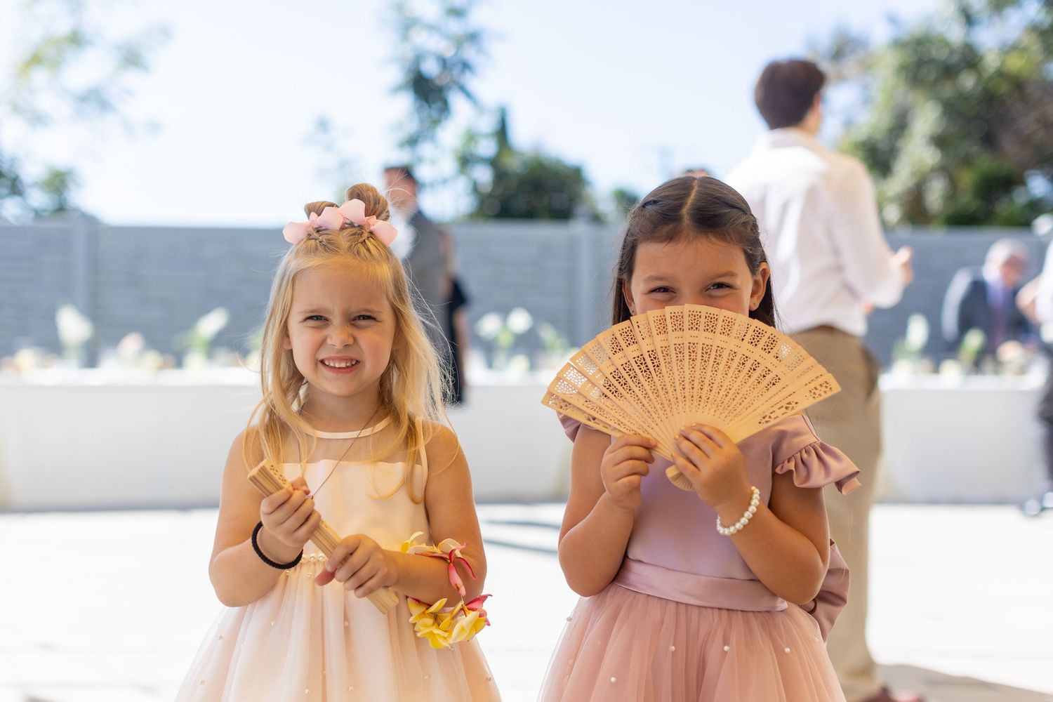 Two young girls in formal dresses hold decorative fans at an outdoor event.