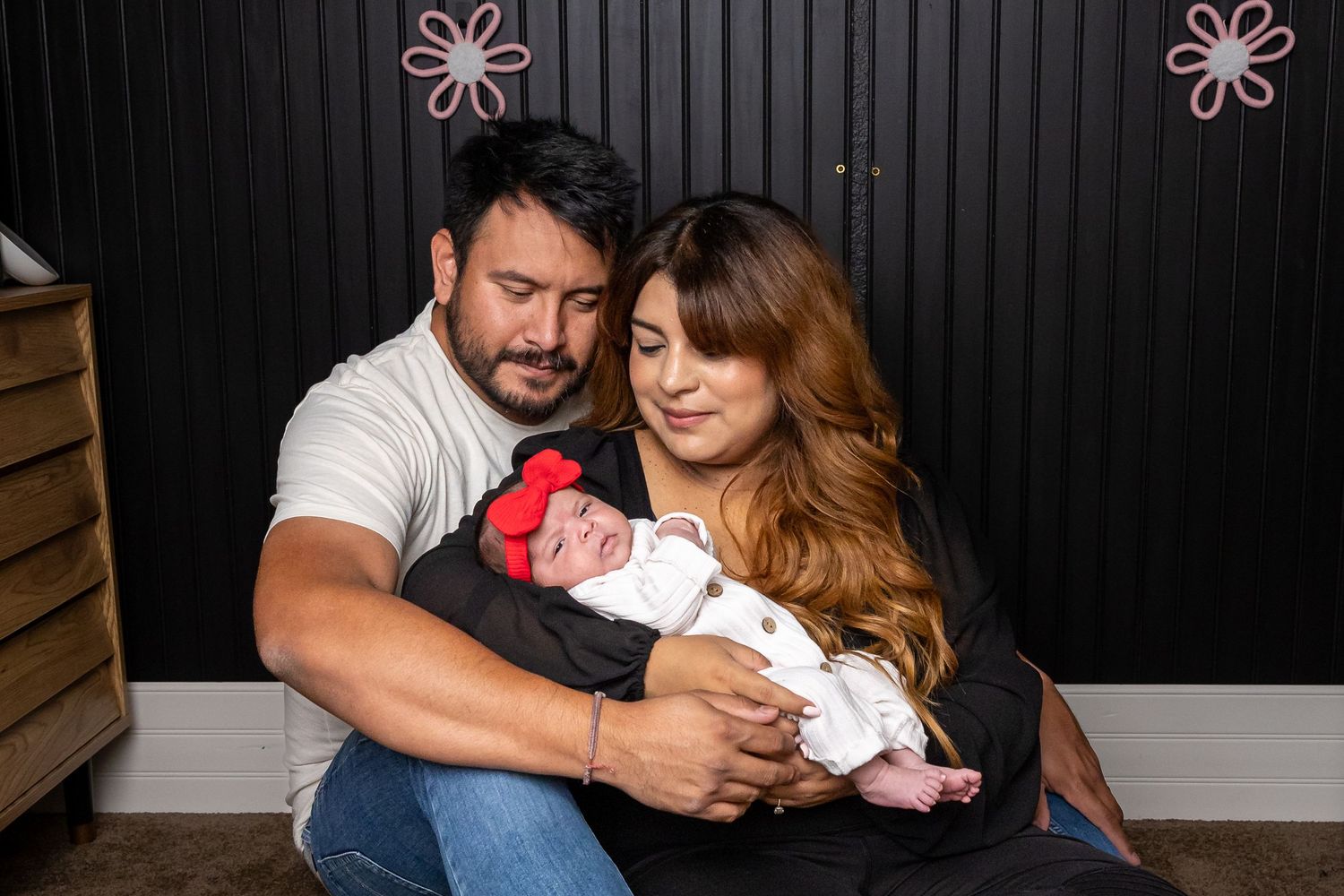 New parents cradling their newborn baby in front of black paneled wall with pink decorations.