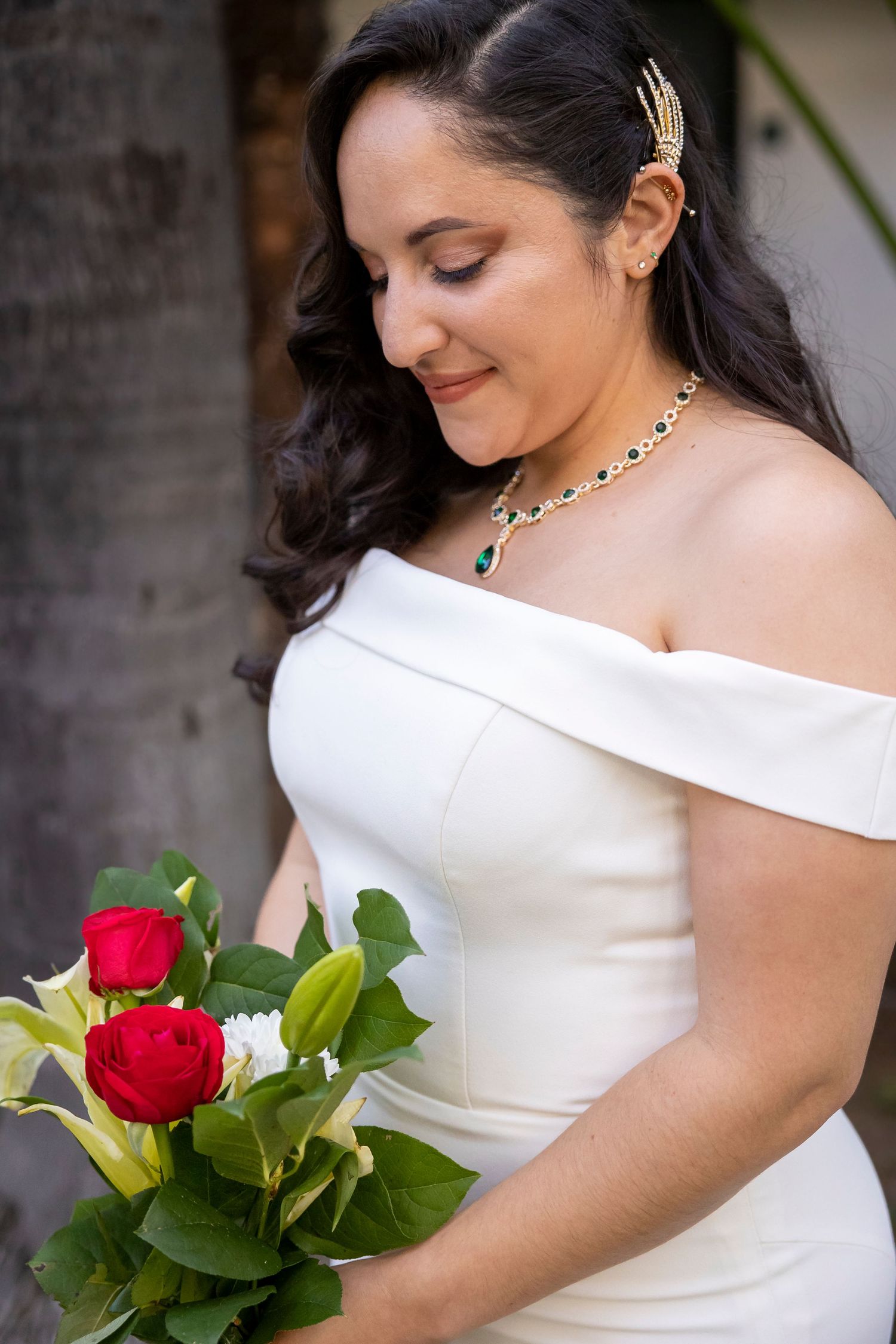 Bride in elegant off-shoulder white dress holds red roses while looking down with a smile.