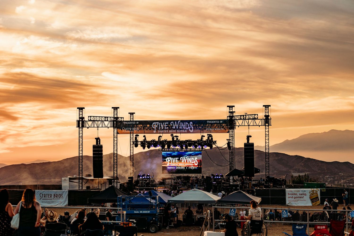 Silhouetted stage equipment and crowd against a dramatic orange sunset at an outdoor concert venue.