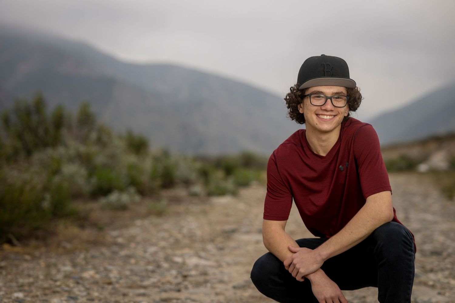 Graduate in burgundy shirt and black hat crouches on desert trail with mountains in background in Yucaipa.