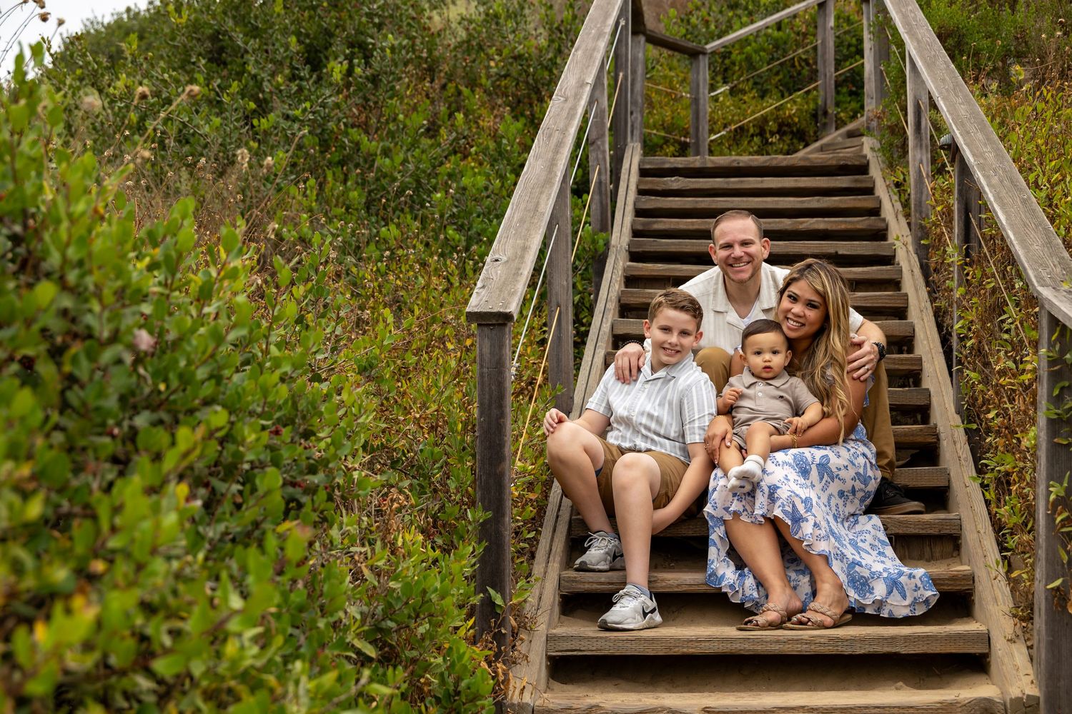 Family sitting together on wooden stairs surrounded by lush green coastal vegetation in Huntington Beach.