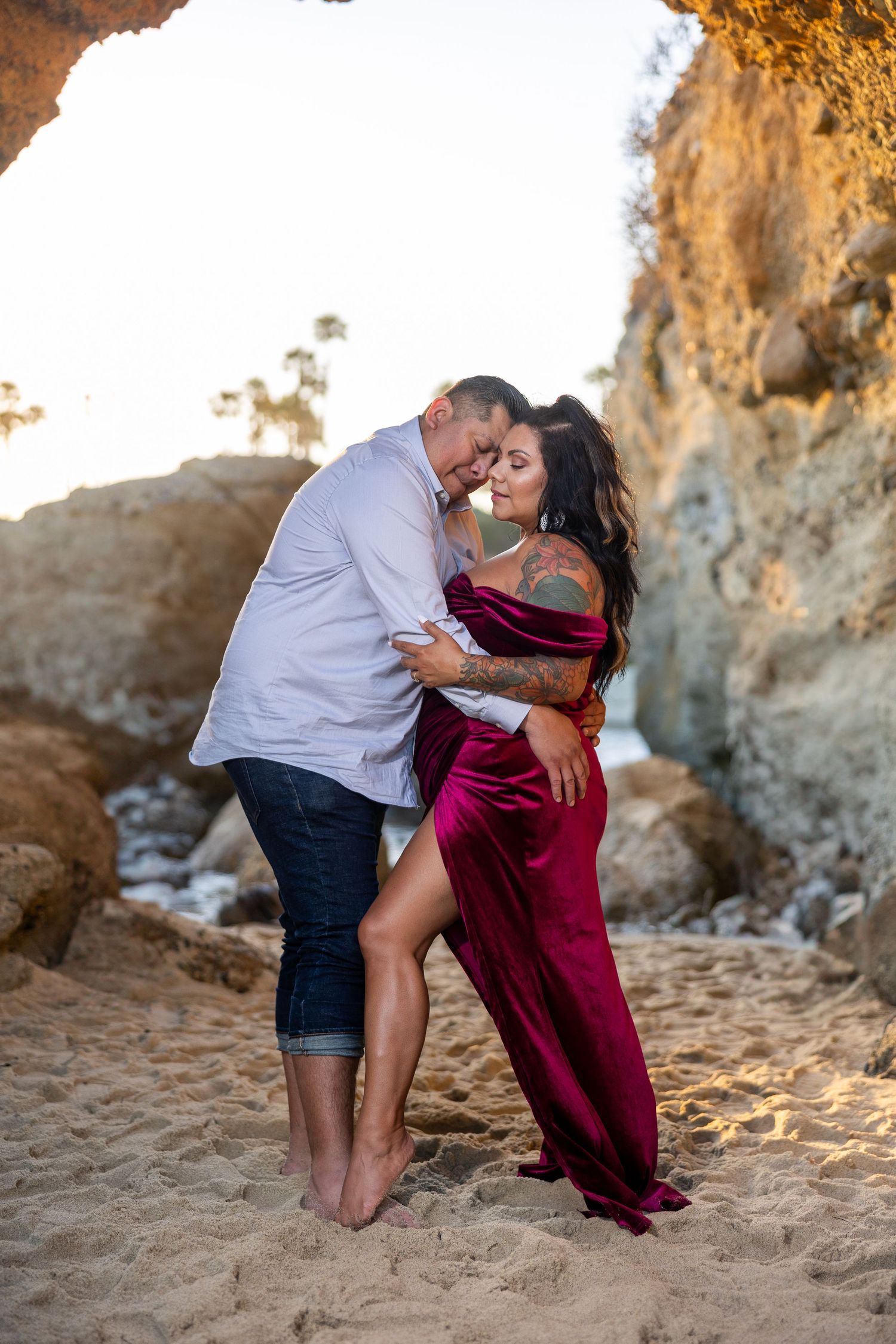 Couple in formal attire posing playfully on beach with cliffs during golden hour.