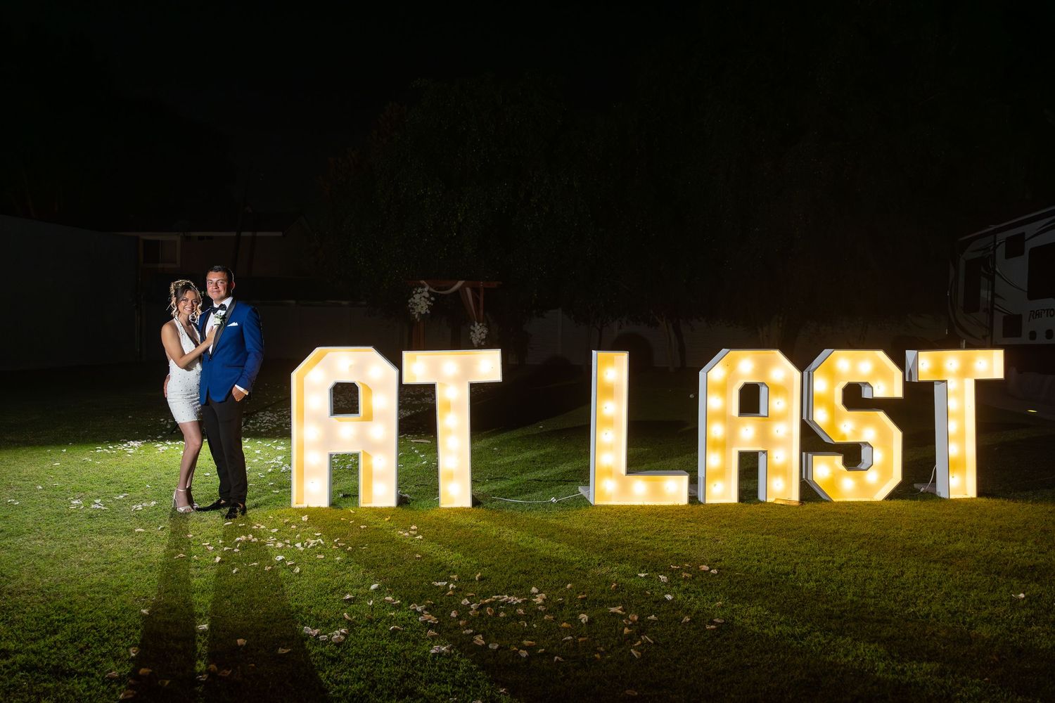 Illuminated AT LAST marquee letters displayed on grass during nighttime wedding celebration.