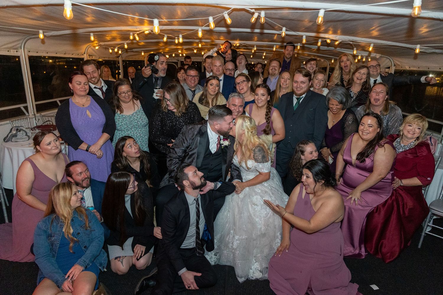 Large wedding party group photo with guests in purple dresses on decorated boat deck.