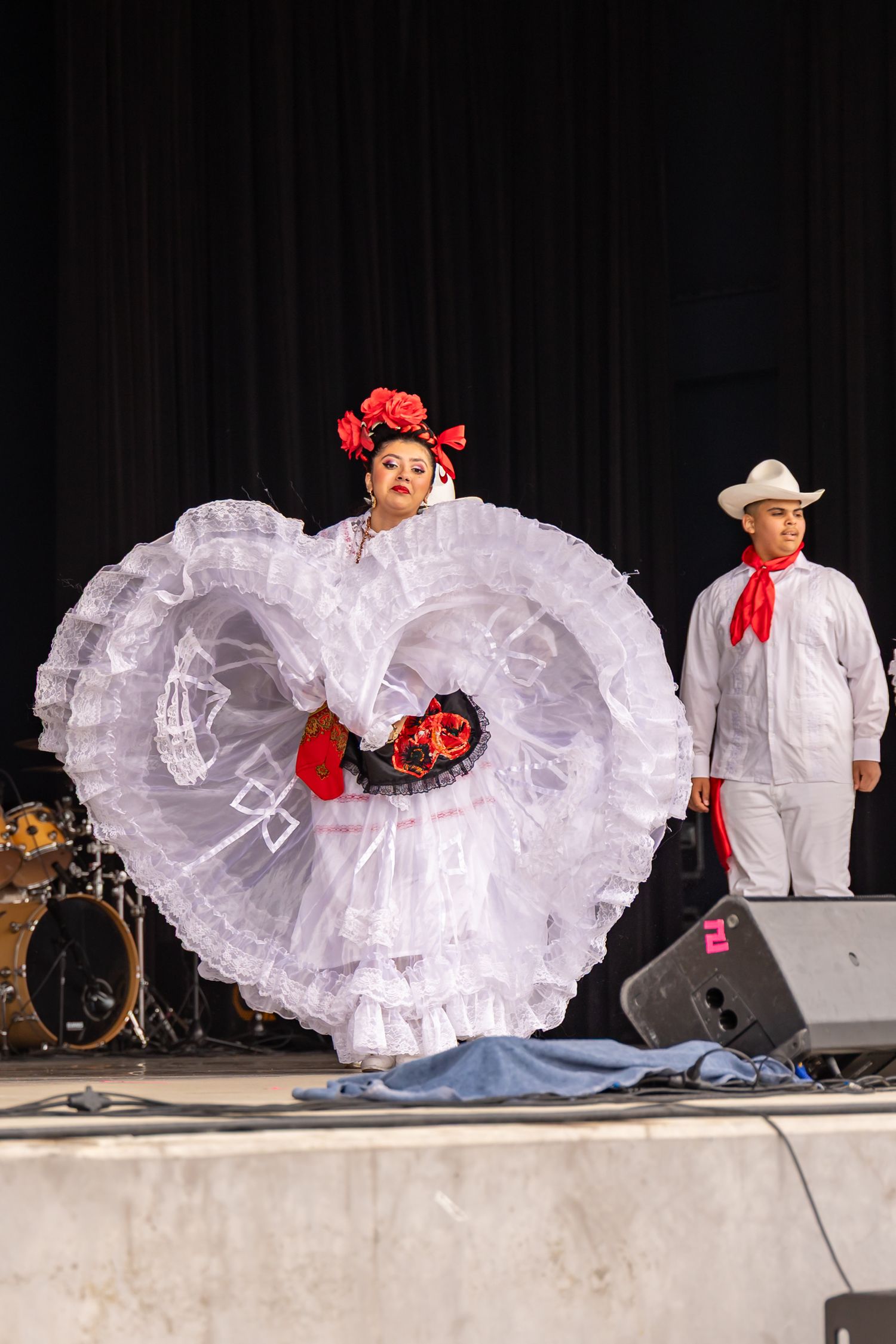 Traditional Mexican folk dancers perform on stage with white flowing dresses and red floral accessories.