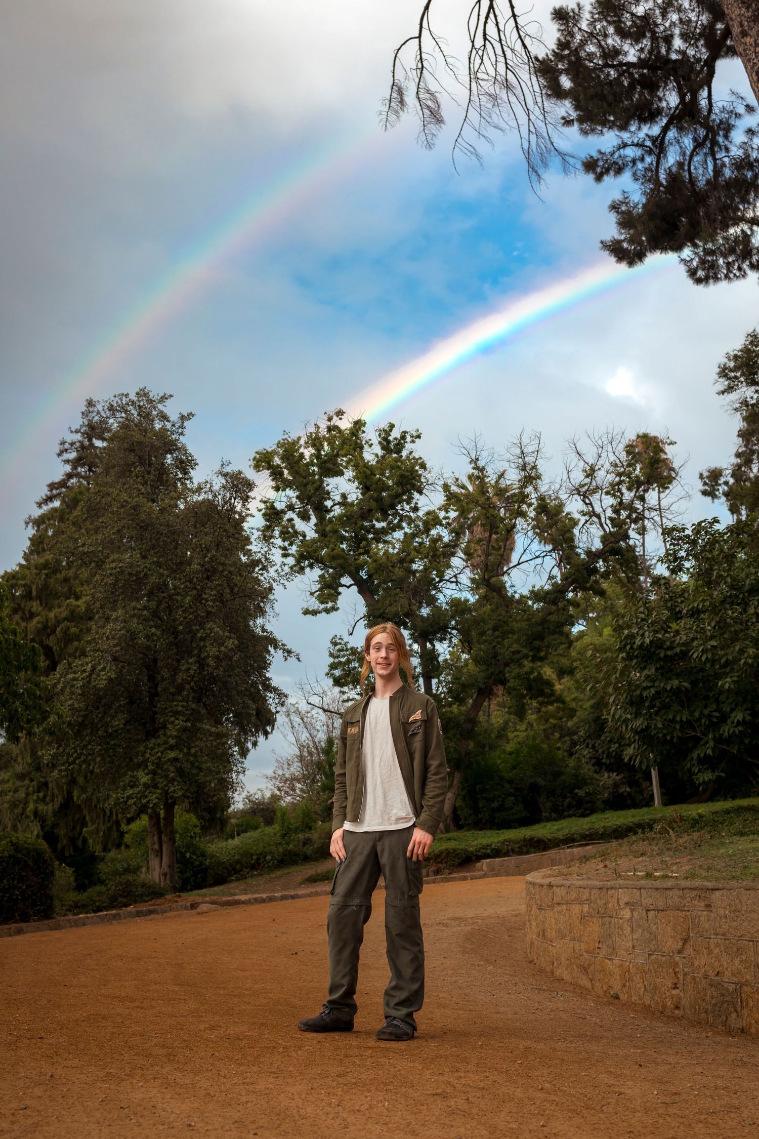 Double rainbow arcs across blue sky above dirt path in wooded area in Redlands.