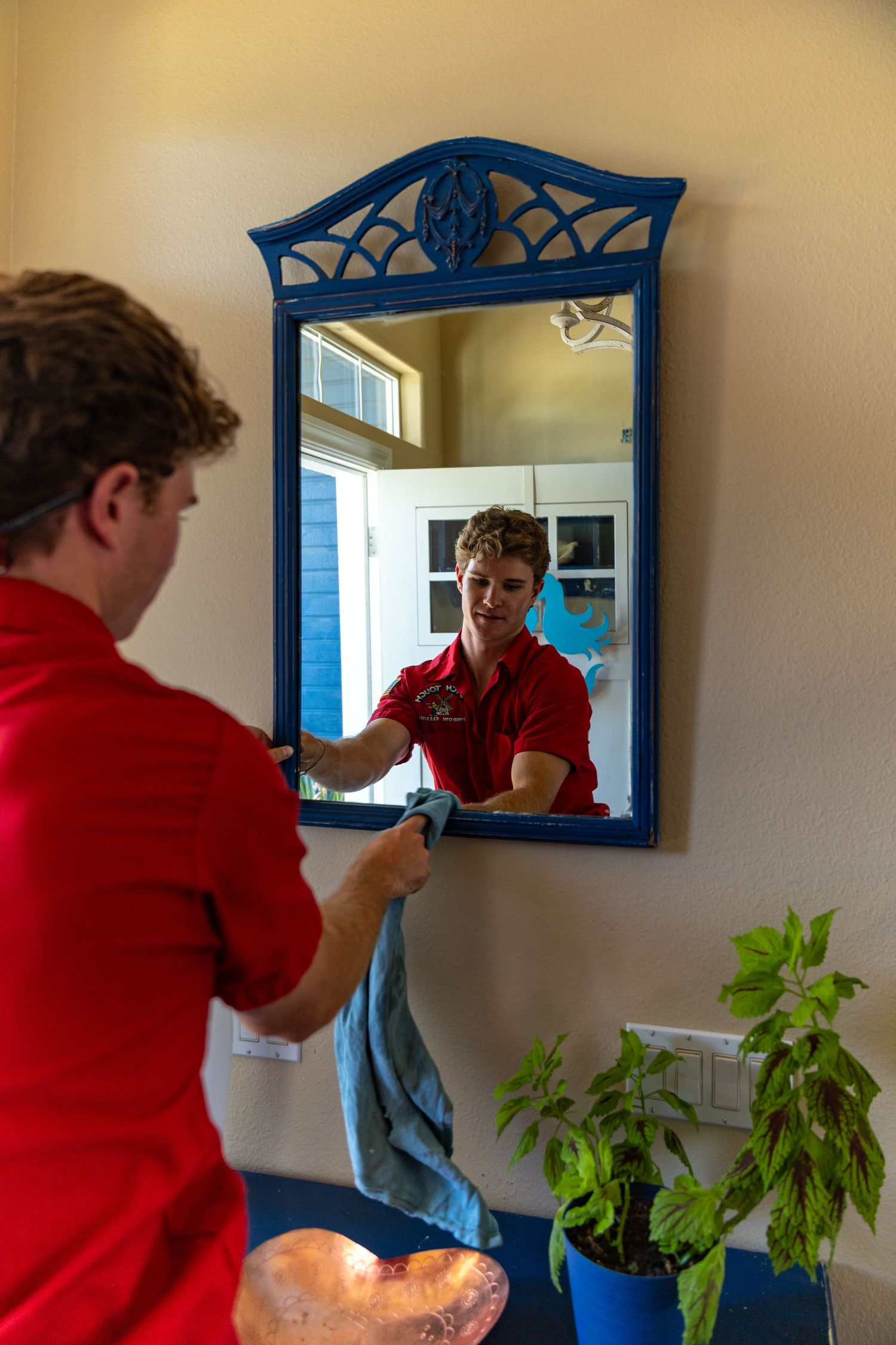 Person in a red shirt adjusts their hair in a bathroom mirror with blue frame and potted plants nearby.