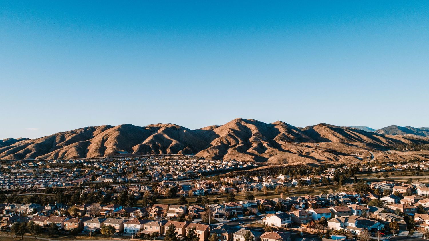 Panoramic view of suburban housing developments and neighborhoods nestled against rolling mountains under blue sky.