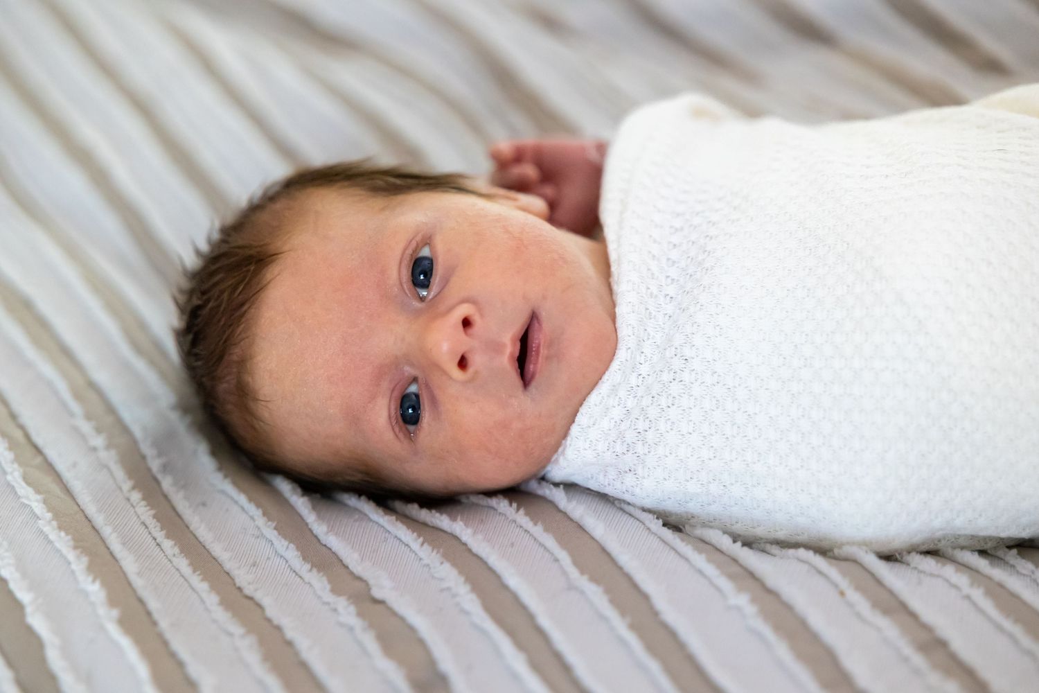 A newborn baby wrapped in white swaddling cloth lies on striped bedding, gazing at the camera with bright eyes in Beaumont.