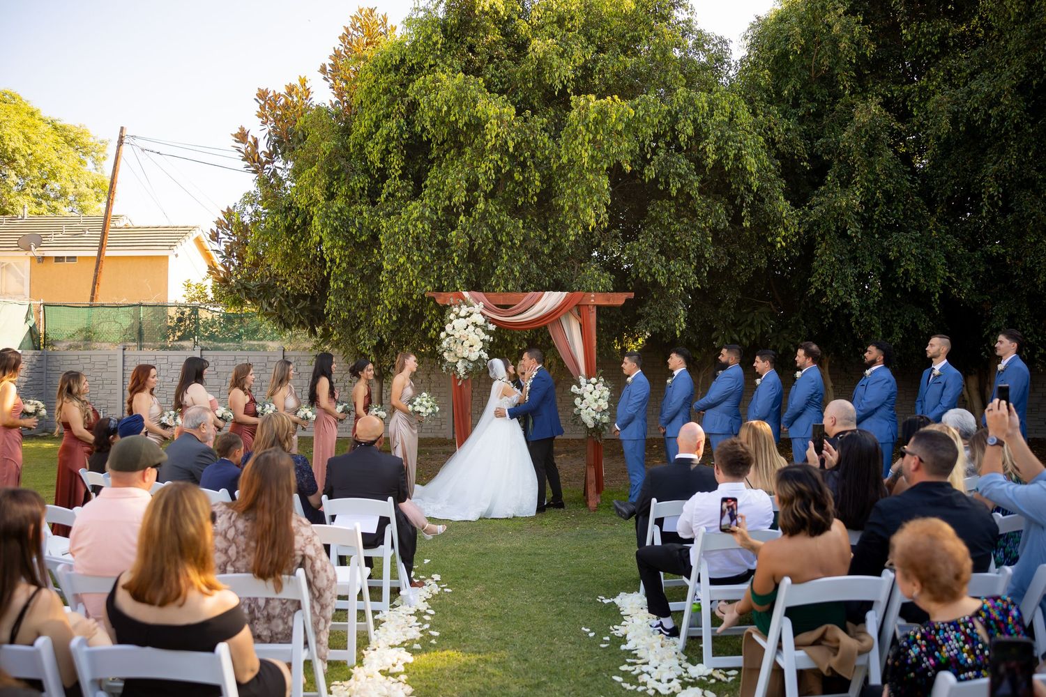 Outdoor wedding ceremony setup with rows of white chairs beneath towering trees.