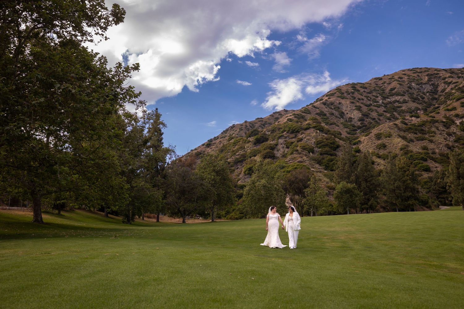 A romantic couple in wedding attire walks across a scenic golf course with mountains in the background.