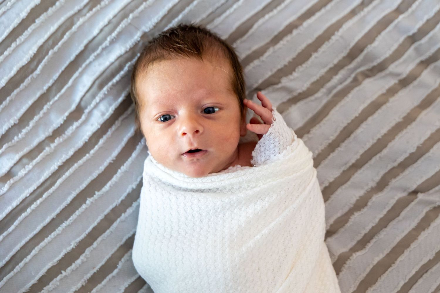 Newborn baby wrapped in white swaddle blanket on striped bedding.