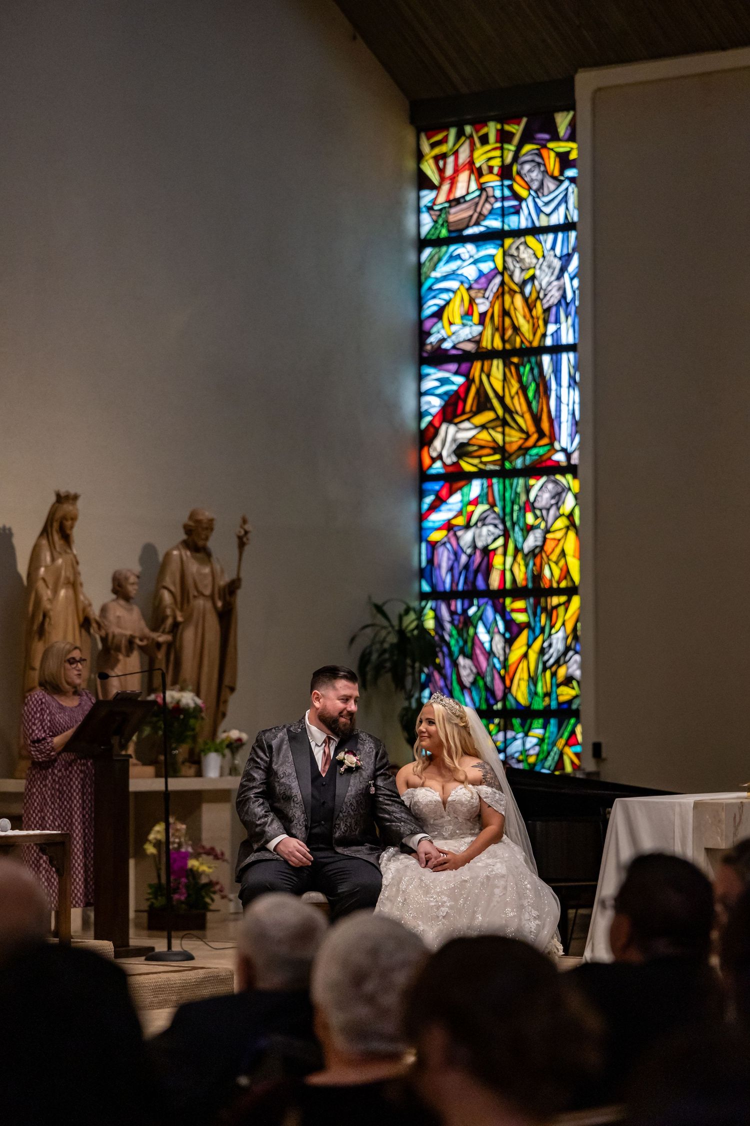 Newlyweds seated in church with beautiful stained glass window and religious statues.