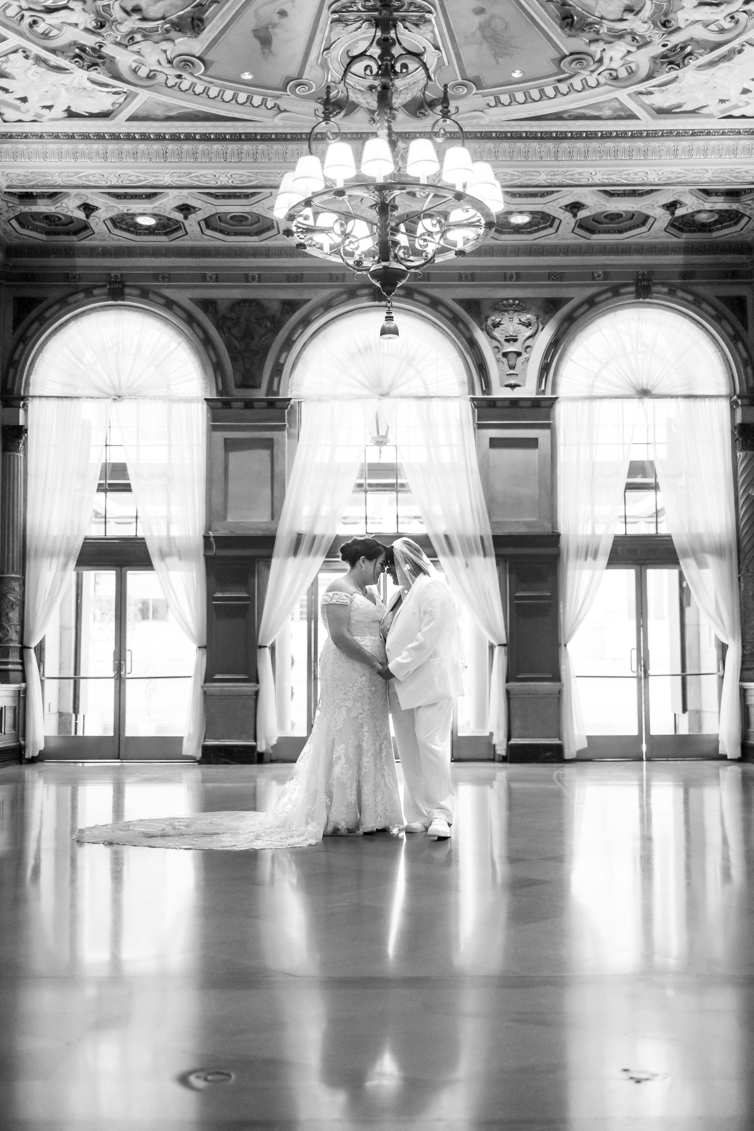 A romantic wedding couple shares an intimate moment in an ornate ballroom with arched windows and chandelier.