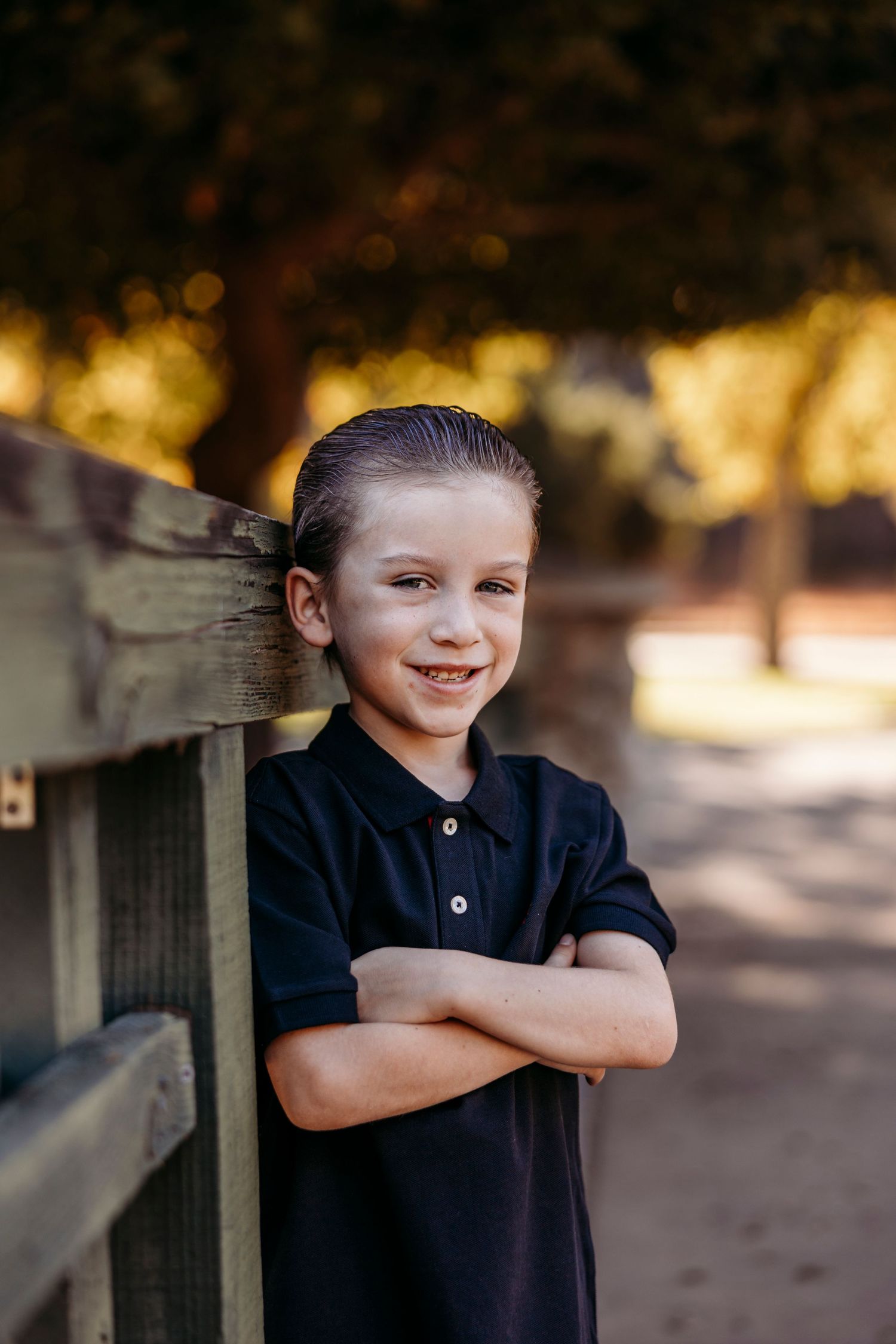 Child in navy blue shirt poses playfully by wooden fence in dappled sunlight.