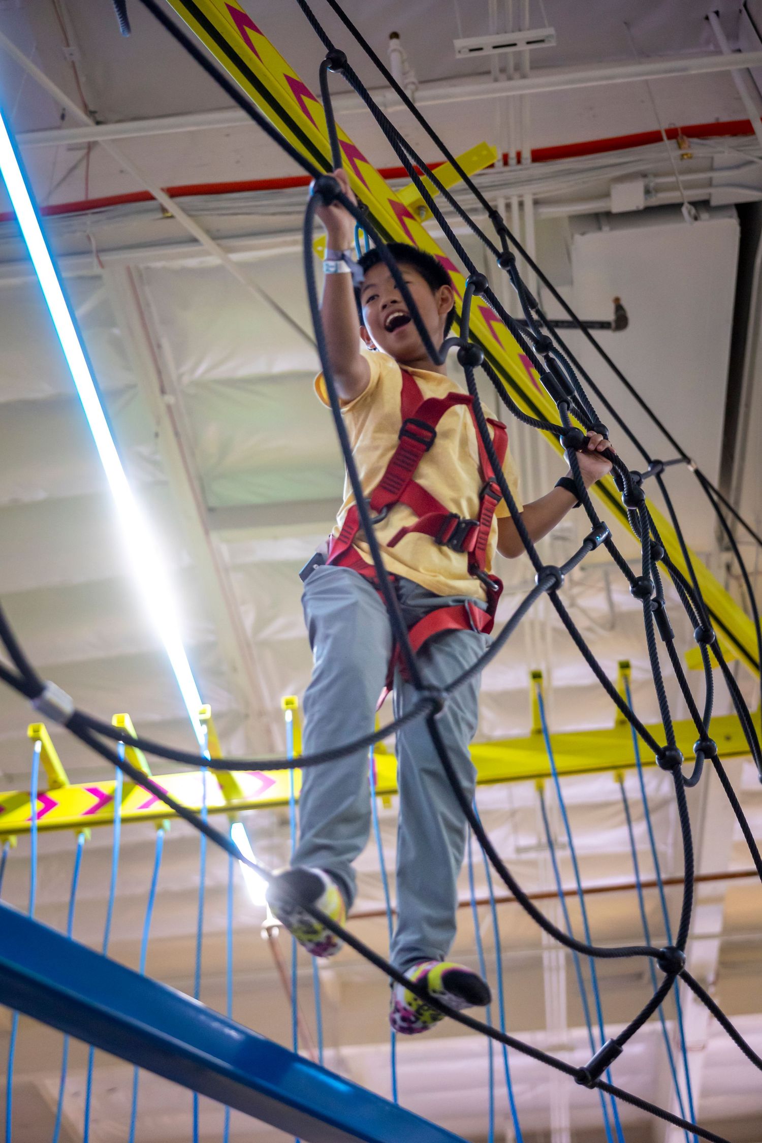 Indoor rope climbing course with bright fluorescent lighting and yellow safety equipment.