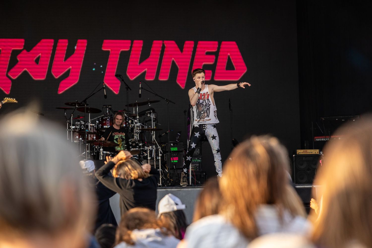 Band performing on stage at outdoor concert with Stay Tuned signage in background.