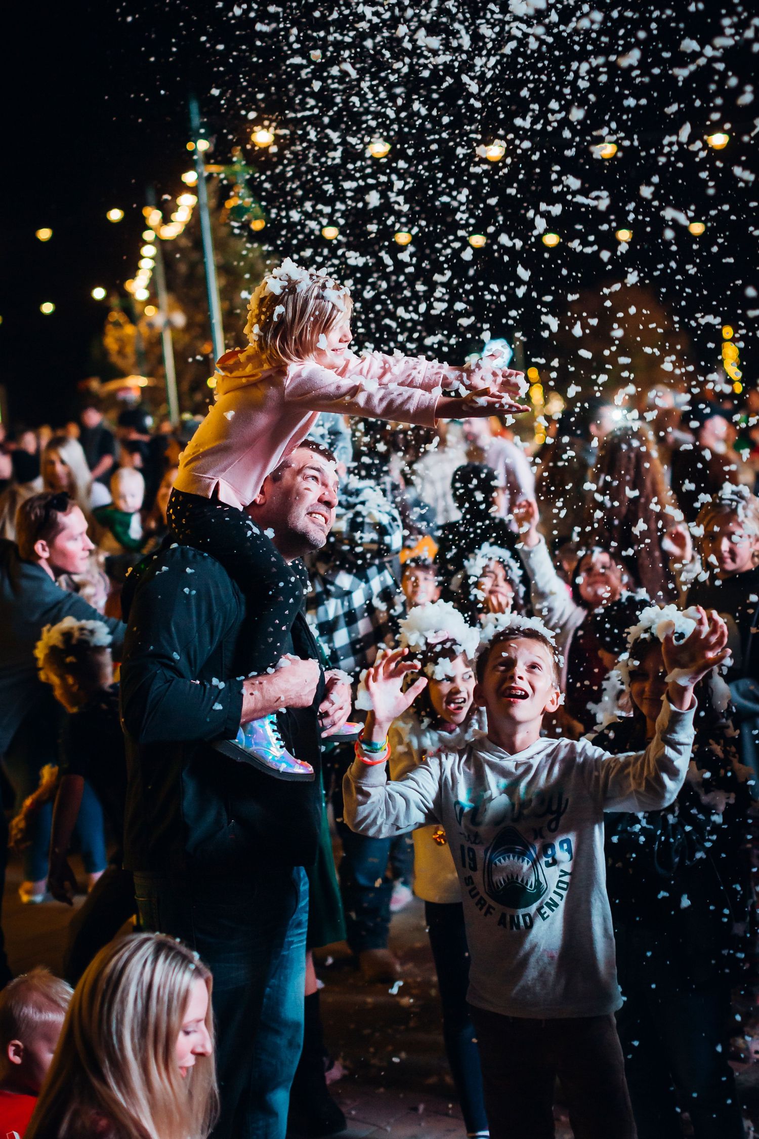 Crowd celebrates at nighttime party with confetti flying through air while string lights glow in background.
