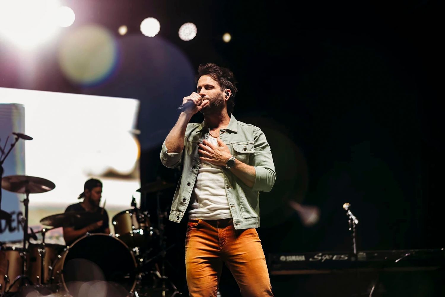 Concert performer singing passionately on stage with dramatic lighting and band setup in the background.