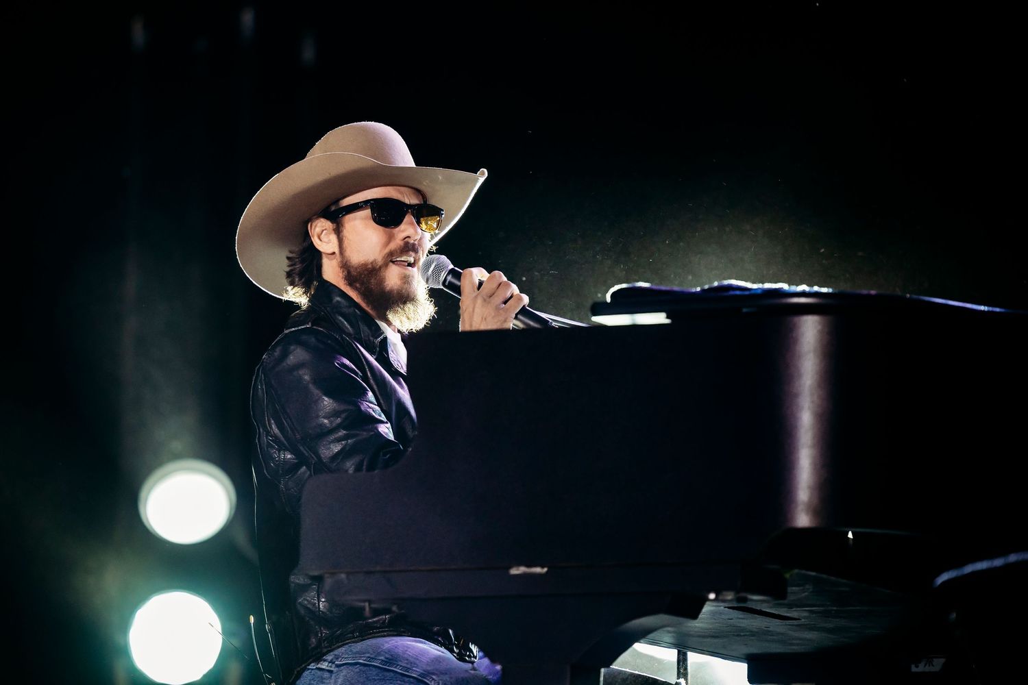 Performer in cowboy hat playing piano on dimly lit concert stage.