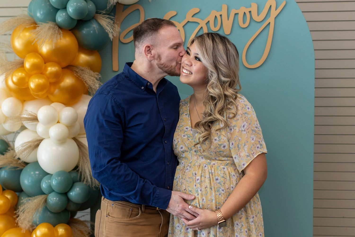 Baby shower celebration setup with teal and gold balloon arch against blue backdrop.
