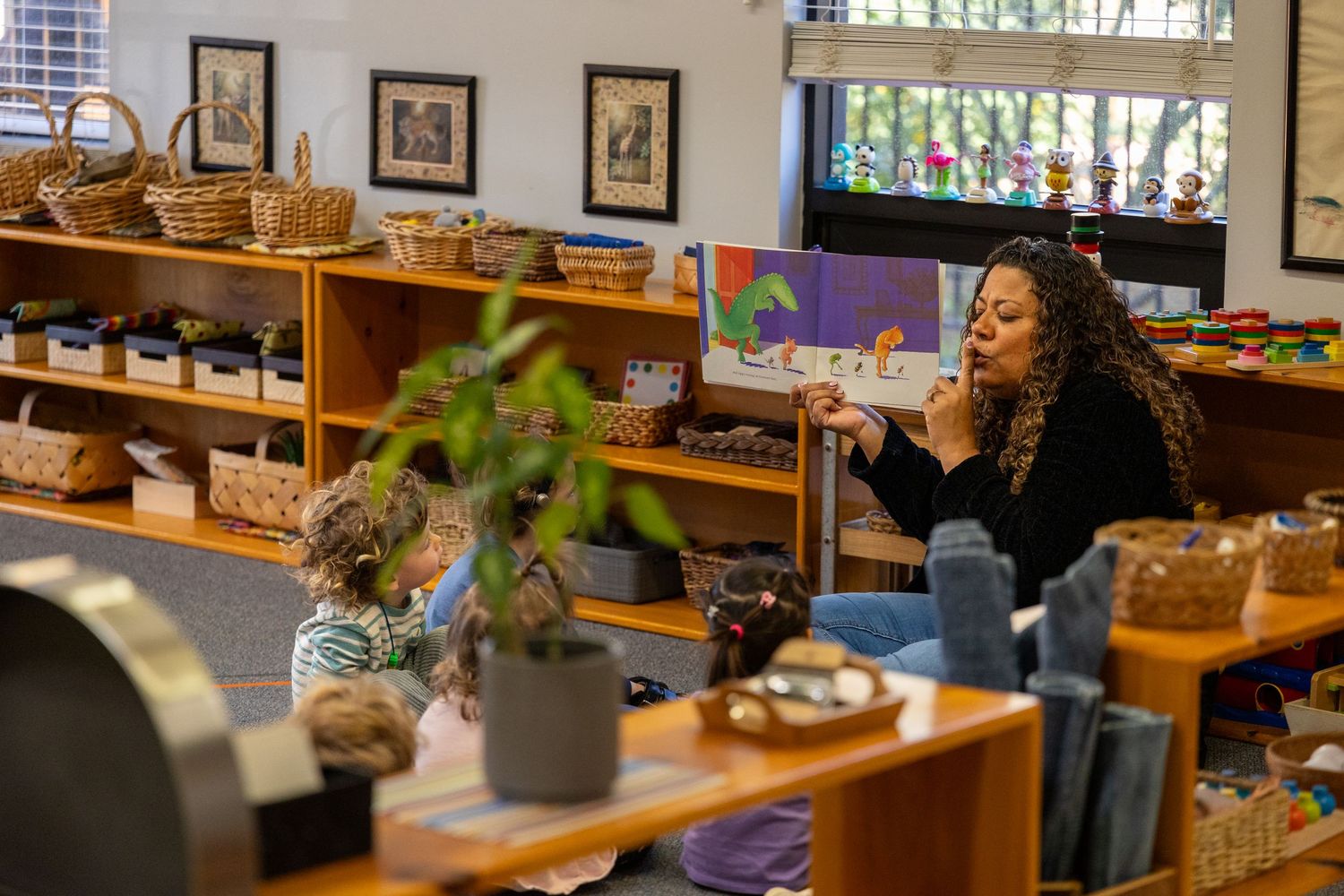 Students engage in a Montessori classroom with wooden materials on shelves and a plant in the foreground.