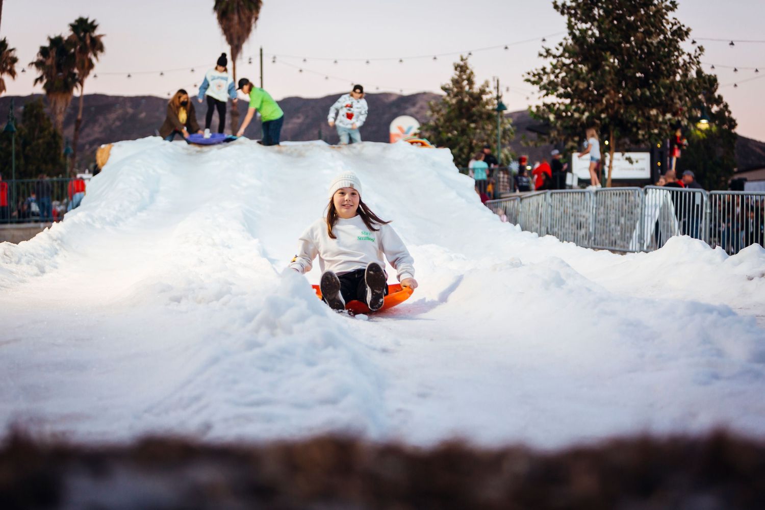Winter sports enthusiasts enjoy snow tubing down groomed slopes at a snowy recreation area with palm trees in view.