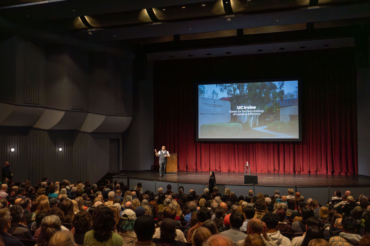 Audience members seated in a dark theater watching multiple screens displaying presentations during a conference event.