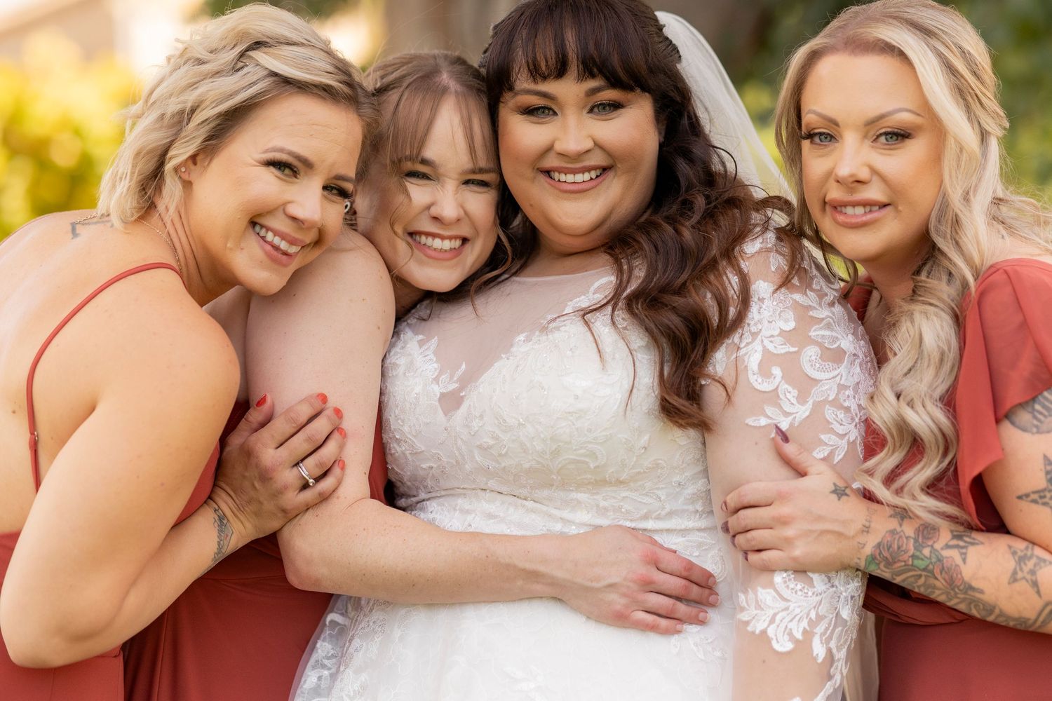 Bridal party in coral dresses smiling and embracing outdoors on wedding day.