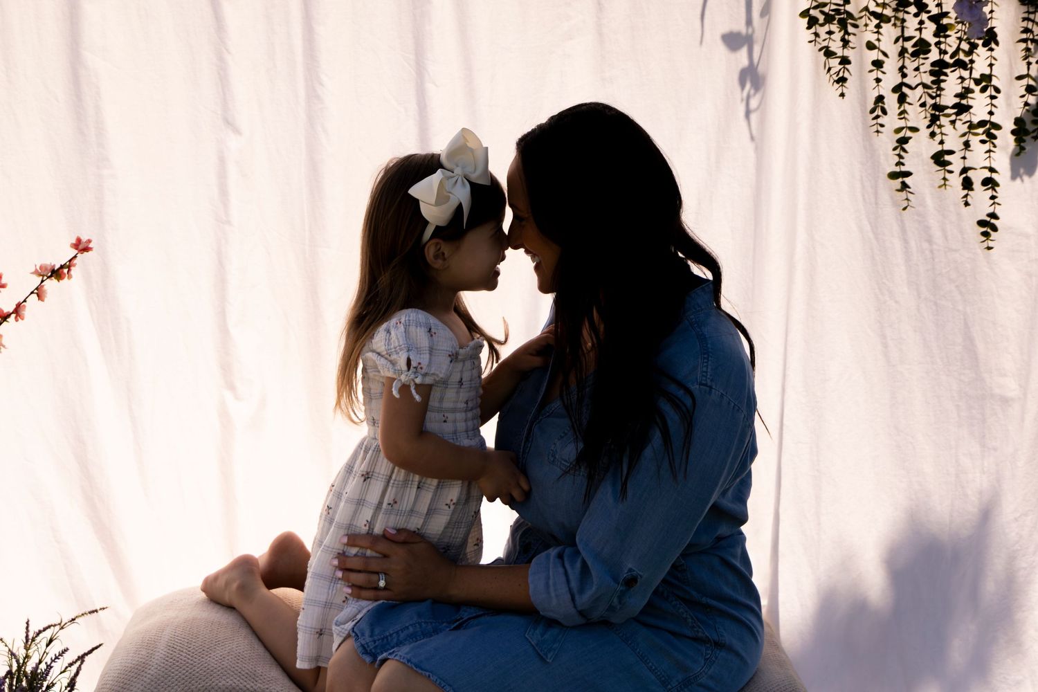 A mother and young daughter share a tender nose-to-nose moment in a softly lit white backdrop portrait.